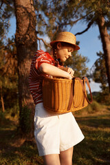 Naklejka premium Young woman outdoors in a straw hat and striped shirt holds a wicker basket, smiling softly as she enjoys a sunny countryside moment. Soft glow highlights leisure.