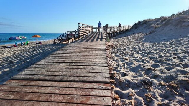 A wide wooden footbridge leading to the beach across a sand dune overlooking the beach, the blue sea and the colorful beach umbrellas on a sunny day.