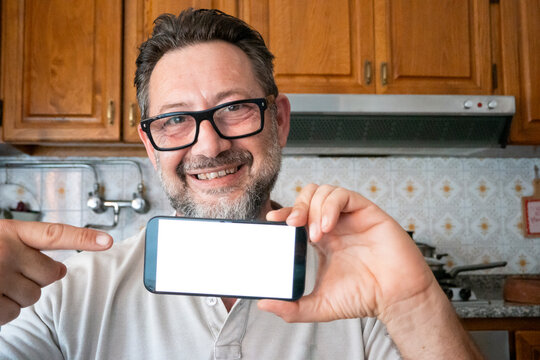 Smiling man in kitchen holds smartphone with blank screen and points to it proudly