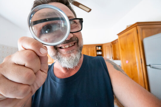 Man smiling in kitchen looking through a magnifying glass at the camera, playful close-up portrait