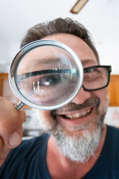 Man with beard smiling and holding magnifying glass up to his eye in casual interior.