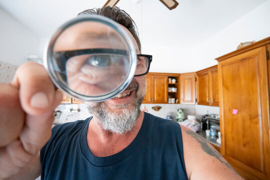 Smiling man holding a magnifying glass in a kitchen, inspecting an item while looking playful