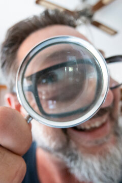 Smiling bearded man holding a magnifying glass in front of his glasses for playful close-up portrait