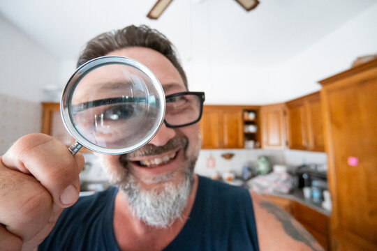Man smiling and holding a magnifying glass to his eye in a bright home kitchen.