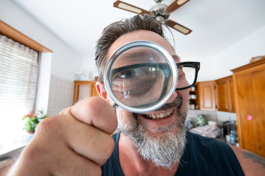 Man smiling in kitchen holds a magnifying glass to his eye for a playful close-up portrait.