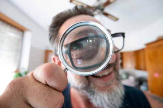 Man holding a magnifying glass to his eye, smiling in a bright home kitchen with a playful, curious expression.