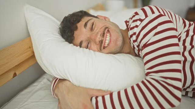 Man hugging a white pillow on bed in building, puckers lips with closed eyes and smiles while wearing red and white striped pyjama and leaning against wooden headboard; morning contentment.