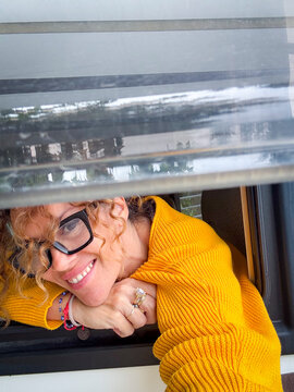 Woman in yellow sweater smiles through a car window, leaning out playfully with glasses