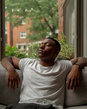 Young Black man relaxing comfortably on sofa by window, eyes closed, taking nap or meditating, enjoying peaceful moment indoors
