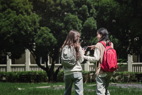 Two Asian university students viewed from behind pointing at smartphone screen together, showcasing genuine friendship and digital communication on green campus lawn during college life