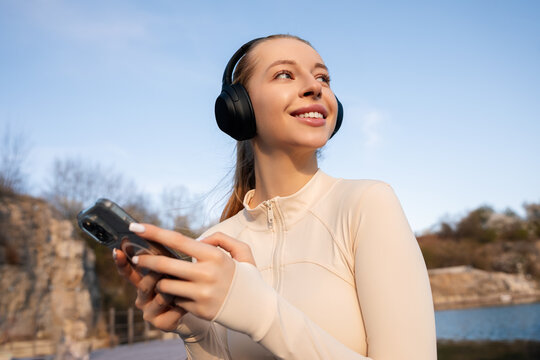 Young woman checking a smartphone outdoors while wearing headphones, smiling with radiant skin and comfortable activewear. Portrait of female model presents modern wellness and digital lifestyle.