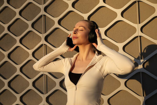 Attractive woman enjoying music with headphones and closed eyes in warm sunlight. Beauty woman portrait combines radiant skin, skincare, activewear and wellness lifestyle concept.