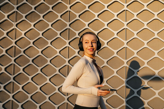 Pretty woman standing with headphones and smartphone in hand, wearing fitted activewear against a geometric wall. Attractive woman presents a stylish fitness and wellness lifestyle.