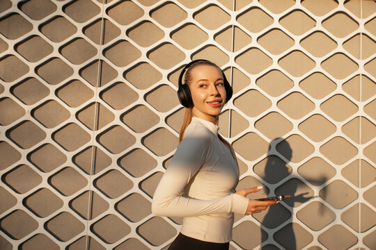 Attractive woman turning with headphones and smartphone in hand, wearing cream activewear under warm sunlight. Young woman shows confident beauty and contemporary wellness lifestyle.