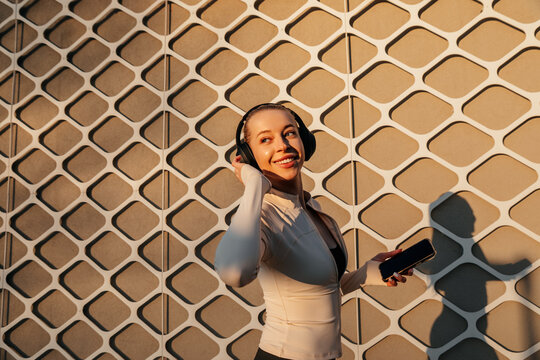 Young woman smiling while adjusting headphones and holding a smartphone, dressed in fitted activewear with warm sunlight. Pretty woman presents a fresh wellness and fitness lifestyle.