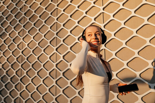 Portrait of female model smiling while listening to headphones and holding a smartphone, wearing modern activewear in golden sunlight. Beautiful woman conveys fitness lifestyle energy.