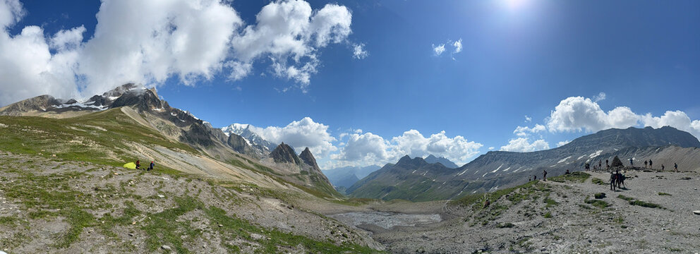 Alpine mountain pass panoramic view rocky peaks glacier green slopes hikers group dirt trail small tarn blue sky white clouds summer landscape wilderness valley