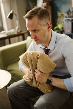 Stressed middle aged Caucasian man squeezing pillow while sitting on sofa, expressing anger and anxiety. Image ideal for mental health awareness and stress management campaigns, vertical