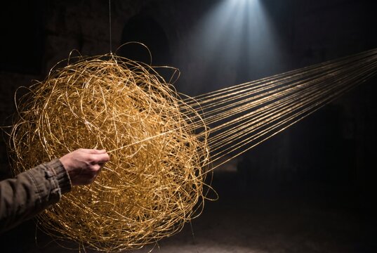 Hand pulling straight threads from massive tangled ball of golden string in dramatic light, problem solving and untangling complexity concept