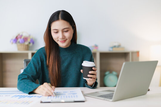 Asian woman smiling, analyzing business reports and financial documents with a pen, holding a coffee cup, sitting at a desk with a laptop, focusing on work from home
