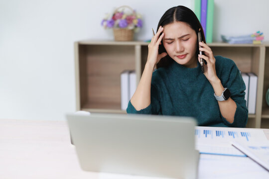 Asian woman experiencing severe headache and stress while working on laptop, talking on phone in office, feeling overwhelmed by demanding business communication and burnout