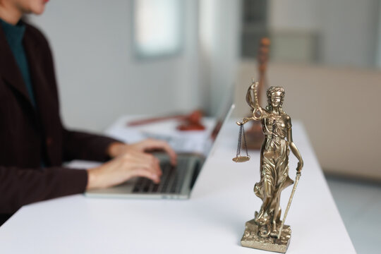 Lady Justice statue symbolizing the legal system and fairness, standing on a white desk with a lawyer working on a laptop in a modern law office setting, representing counsel and advocacy