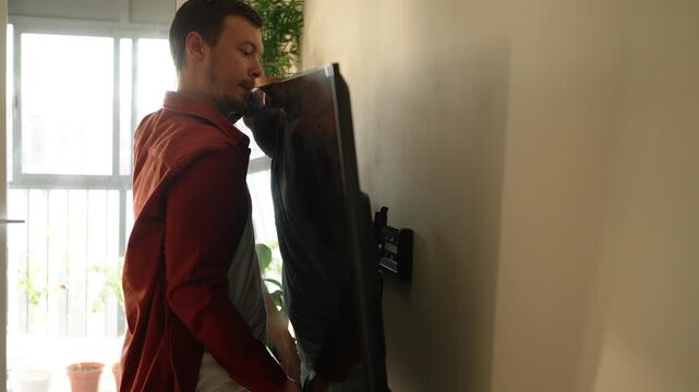 Focused man carefully mounting a brand new flat screen tv onto the wall bracket in his modern apartment, performing a do it yourself home renovation and setting up the electronic device