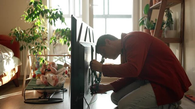 Dedicated father multitasking at home, carefully assembling and mounting a new television on the wall while his young child watches from a nearby bouncer in their bright, contemporary apartment