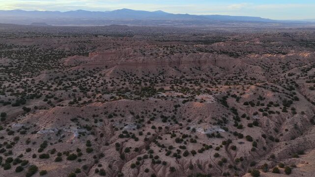4K aerial view over the eroded sandstone formations of the Nambe Badlands in New Mexico. Scattered juniper trees dot the deeply carved, arid clay landscape under a clear sky.