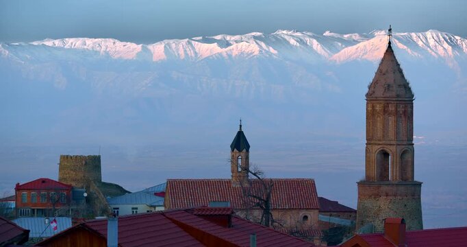 Historic Georgian church architecture rises above layered red metal and clay tiled roofs with tall brick bell towers fortress masonry and the vast snow covered Caucasus range behind the old town