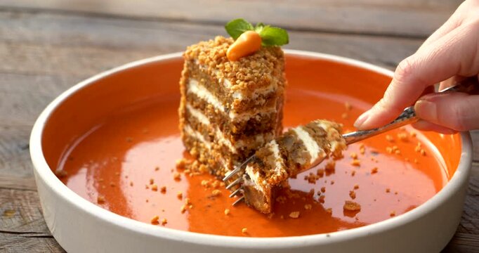 Hand presses a fork into a tall layered carrot cake then lifts a tender bite revealing alternating moist sponge and cream filling beneath nut crumbs mint leaves and a small decorative carrot