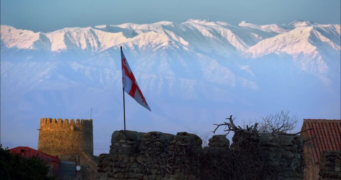 Close Georgian flag flutters and folds above a rugged old stone rampart with a square medieval tower and red tiled roof framed against luminous snow covered Caucasus ridges in soft winter light