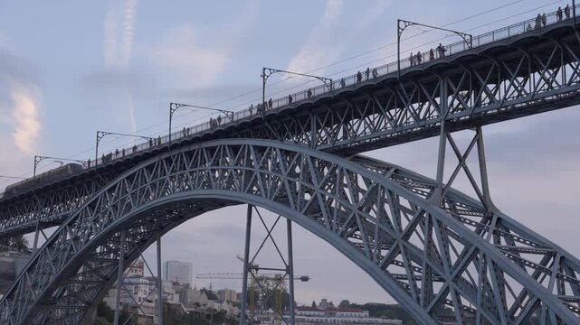 Static upward tilt towards the Dom Lu&iacute;s I bridge as an electric metro train crosses and pedestrians walk along the upper deck at sunrise. Shot in 4K at 25 fps.