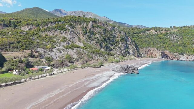 Aerial drone view flying over Limnionas Beach, revealing vibrant turquoise waters, rugged coastal cliffs, lush Mediterranean nature, and a colorful sky in Evia, Greece.