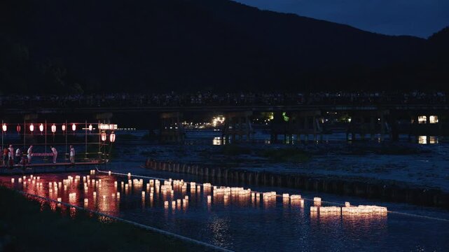 Floating Paper Lanterns Glow on River at Night with Distant Bridge Crowd