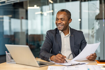 Obraz premium Experienced entrepreneur smiling while reviewing financial paperwork and signing contracts at his desk, managing business operations with a laptop in a modern corporate workspace