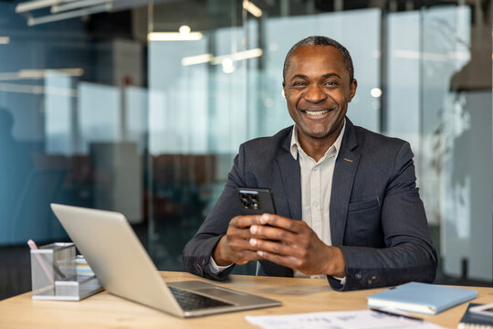Mature businessman sitting at an office desk. Smiling at the camera while holding a smartphone. With a laptop and documents nearby