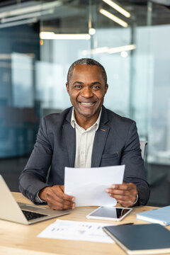 Mature businessman smiling at desk, reviewing paperwork with laptop and tablet, actively managing business tasks in a contemporary office setting, representing professional success and leadership