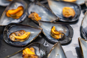 Fresh steamed mussels resting on open blue shells with tender orange meat visible, displayed on a wet dark surface at a coastal market, a concept of sustainable fishing trade. © Masarik