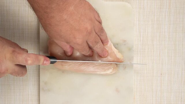 A raw chicken breast is being sliced lengthwise with a kitchen knife on a white cutting board. A long strip of chicken is separated beside the main piece.