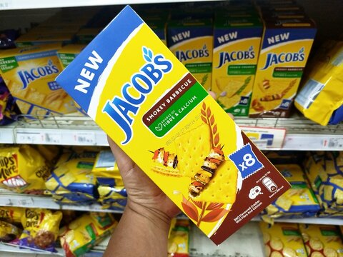 KUALA TERENGGANU, MALAYSIA- AUGUST 20. 2026: Close-up of a hand holding Jacob's Smokey Barbecue crackers packaging inside a hypermarket aisle, with stacked biscuit boxes on store shelves.