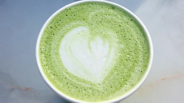 Top down close up view of fresh matcha latte with white heart shaped milk foam art in takeaway cup.