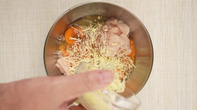 Grated cheese pours from a glass cup into a bowl of cracked eggs and peppered chicken. This close-up shows a homemade batter assembling.