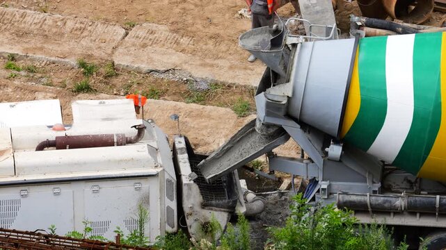 A concrete mixer truck discharges fresh liquid cement through a chute into a white construction pump machine on a dusty daytime building site for a real estate project.