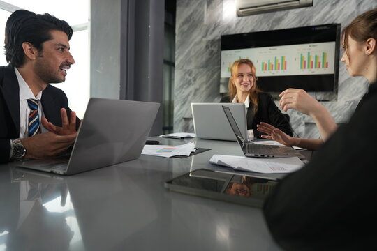 Manager laughs with palms apart as the young colleague holds her gesture, humor loosening the morning meeting in the glass walled office in a bright modern coworking office.