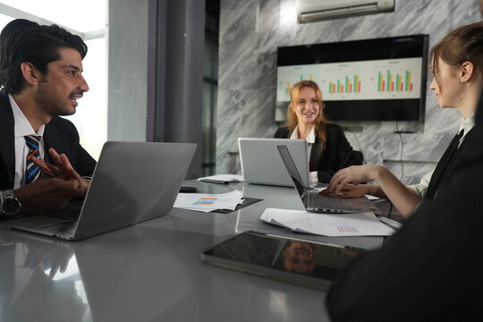 Fingers raised as he counts his points, the dark haired manager briefs the two women across the sunlit boardroom table by the windows in a bright modern coworking office.