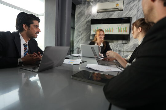 Both hands raised in a wrapping up gesture, the manager closes the morning briefing while his two colleagues smile over their laptops in a bright modern coworking office.