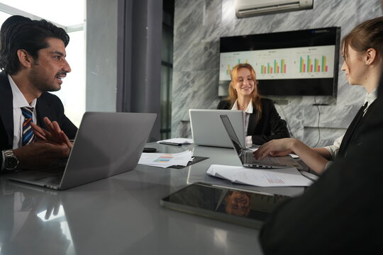 Hands opened mid sentence, the manager talks through the plan while the blonde teammate attends from the television end of the bright table in a bright modern coworking office.