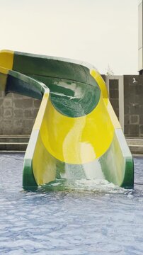 A colourful children's water playground (splash park) located in an outdoor swimming pool, situated on a rooftop setting with a modern glass-walled building in the background.