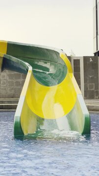 A colourful children's water playground (splash park) located in an outdoor swimming pool, situated on a rooftop setting with a modern glass-walled building in the background.